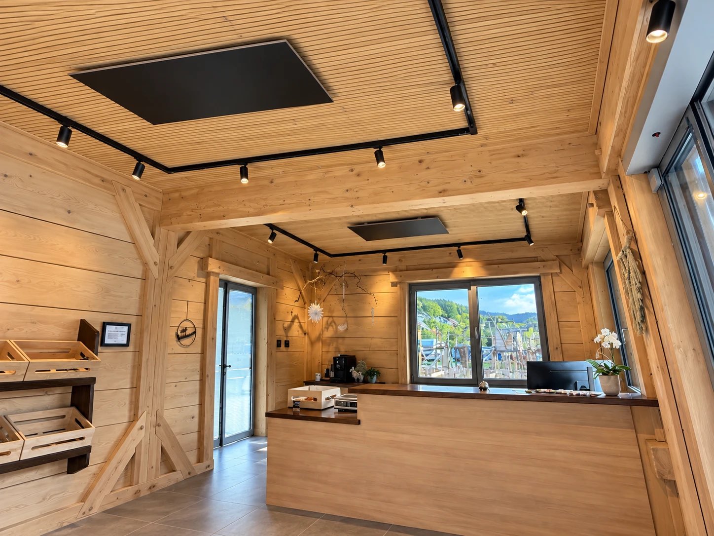 Empfangsraum in Holzbauweise mit zwei dunklen Infrarot-Deckenpaneelen in der Holzdecke, Empfangstheke und Fenster zum Campingplatz.