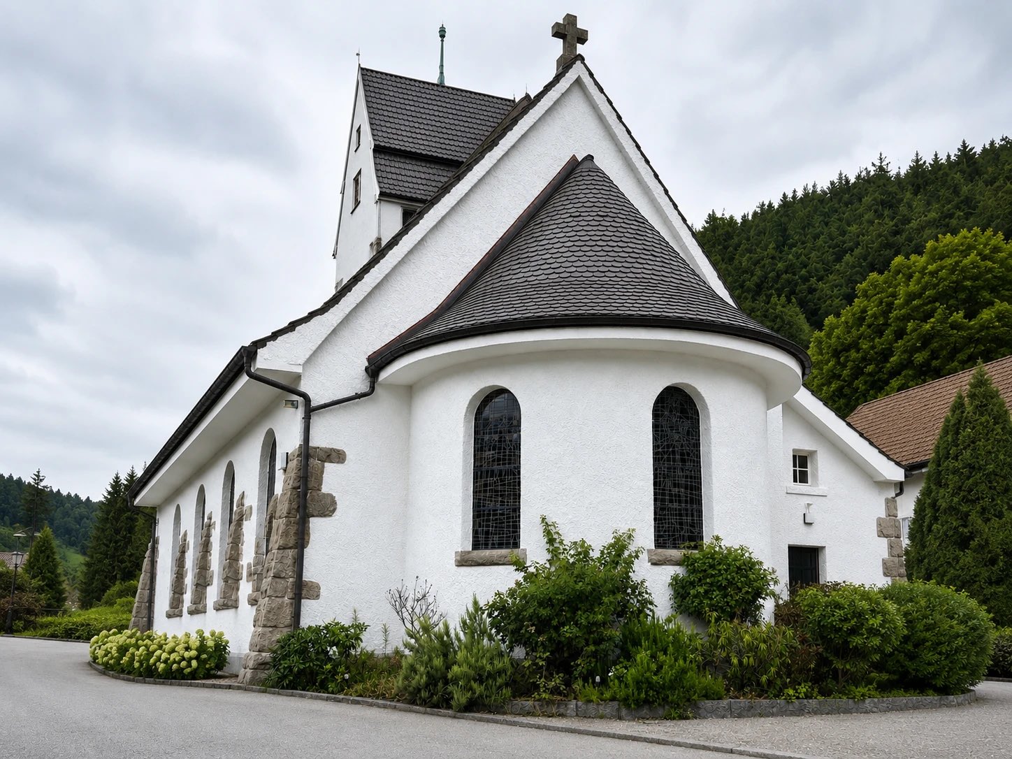 Weiß verputzte Kirche mit Turm, Schieferdach und hohen Rundbogenfenstern vor bewaldetem Hang.