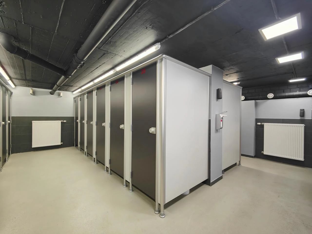 WC-Trennwandanlage mit anthrazitfarbenen Türen und hellgrauen Wangen in einem Sanitärraum mit Sichtbetondecke.